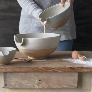 Person pouring liquid from a ceramic bowl into another on a wooden table.