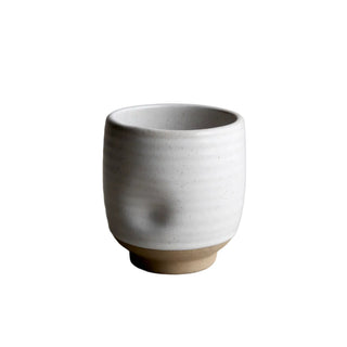 A handmade ceramic cup with a textured, ribbed white glaze and a natural, unglazed tan base, set against a neutral gray concrete background.