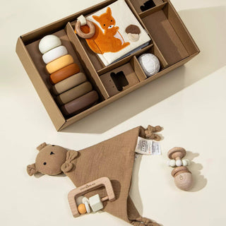 Baby toy set with wooden rings, teething ring, and soft blanket in a box on a light background