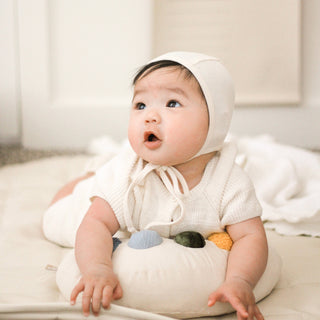 Baby wearing a white hooded outfit sitting on a cushion with toys.