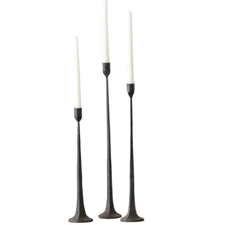 Three black candlesticks with white candles on a wooden surface against a white wall.