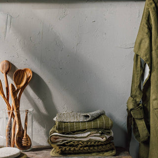 Wooden spoons in a glass jar on a wooden surface with green towels and a green jacket against a white wall.