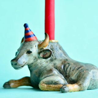 Ceramic bull figurine with a party hat holding red candles against a light blue background