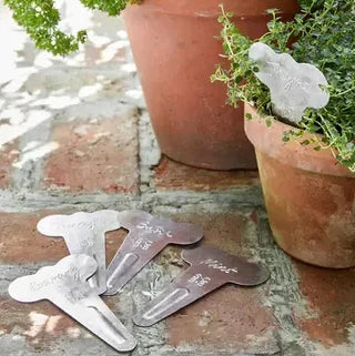 Metal plant markers with engraved names next to potted plants on a brick surface