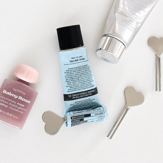 A styled flat-lay of various cosmetic products on a white background, including a light blue squeeze tube of hair oil, a pink bottle of lip gloss, a silver tube, and silver heart-shaped tools.
