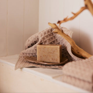 Bar of soap on a wooden tray with a towel and branch against a tiled wall.