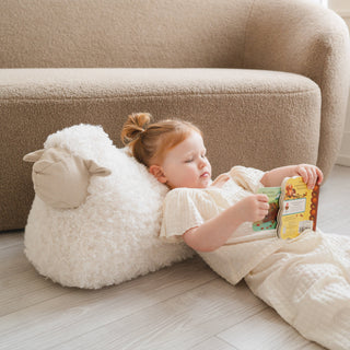Child lying on a fluffy white pillow with cartoon face, reading a book in a cozy living room.
