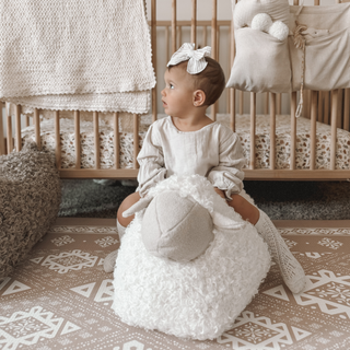 Baby sitting on a textured stool in a nursery with wooden crib and decorative pillows.