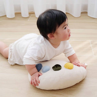 Baby sitting on a cloud-shaped pillow with colorful buttons on a wooden floor.