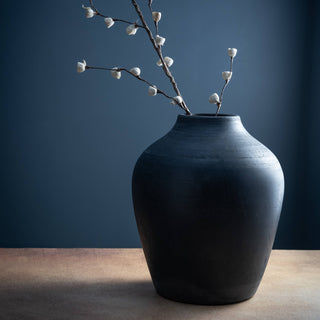Black vase with dried branches on a wooden surface against a dark blue wall