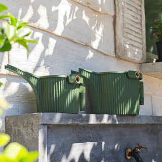 Two green compost bins on a stone surface with a building and plants in the background.