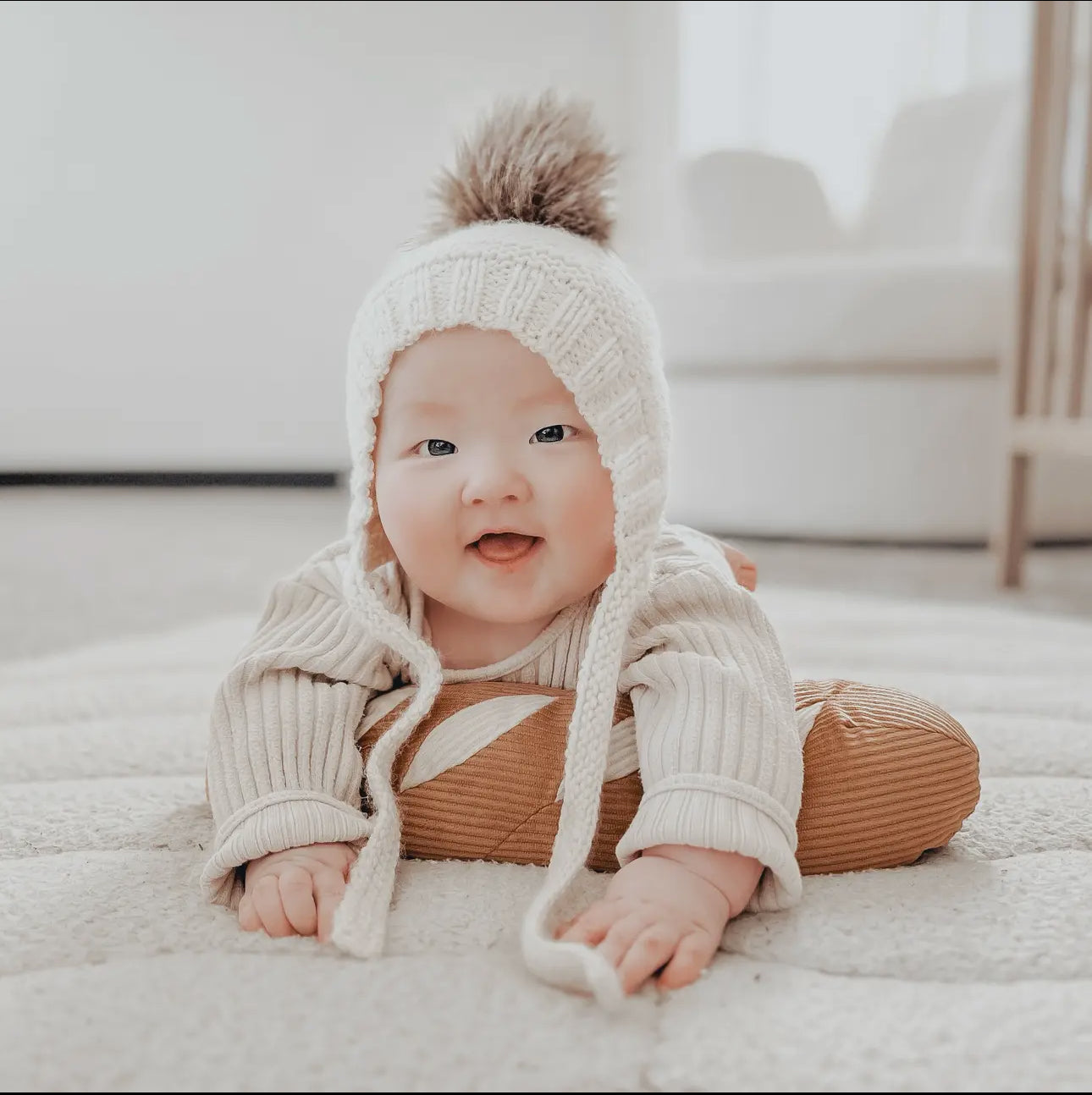 Baby wearing a knitted hat with a fluffy pom-pom, laying on a bread pillow.