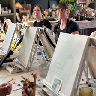 Two women sitting at a table with art supplies, including easels and paintbrushes, in a studio setting.