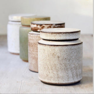 Four handmade ceramic canisters with matching lids, arranged in a diagonal row. The canisters feature a speckled glaze in various earthy tones including cream, beige, light green, and brown, set against a neutral, softly lit background.