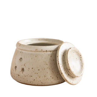 A speckled ceramic lidded container with a matching lid leaning against it, photographed on a warm beige studio background.