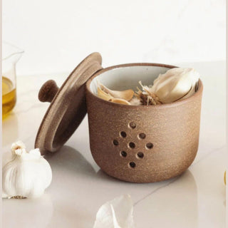 A brown speckled ceramic garlic press container with its lid off, showing garlic cloves inside, set on a light marble surface with a garlic bulb nearby.