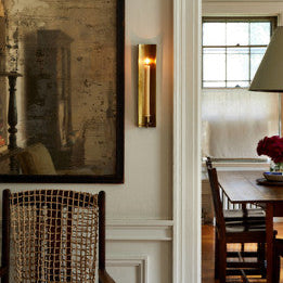A brass plated channel wall sconce mounted on a white wall, with a warm glow emitting from it, reflecting on the wall above a mirrored art piece and a window.