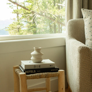 Small wooden stool with books and a vase next to a sofa by a window with a view of trees.