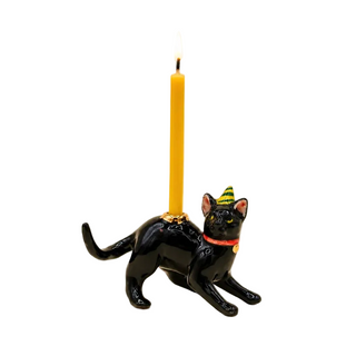 A black cat-shaped candle holder with a lit yellow candle inserted into its back. The cat is wearing a red collar and a green and yellow party hat, set against a plain beige background.