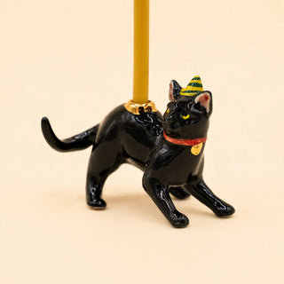 A glossy black cat figurine wearing a yellow and green striped party hat and a red collar with a gold heart, holding a gold pole against a warm beige background.