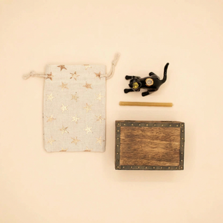 A flat-lay product shot featuring a light gray fabric drawstring pouch with metallic gold star patterns, a small black cat figurine with a gold medallion, a wooden stick, and a dark brown wooden box with metal studs and an embossed logo, all arranged on a solid cream background.