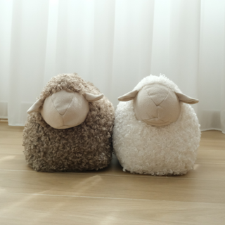 Two fluffy sheep-shaped cushions, one brown and one white, on a wooden floor with a white curtain background.