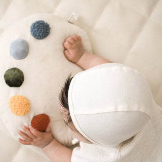 Baby lying on a soft surface with colorful pom-poms, wearing a white hat.
