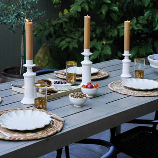 Outdoor dining table set with candles, plates, and glasses against a green foliage background