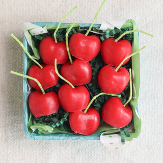 A box of red cherry-shaped birthday candles with green stems, arranged in a blue berry basket.