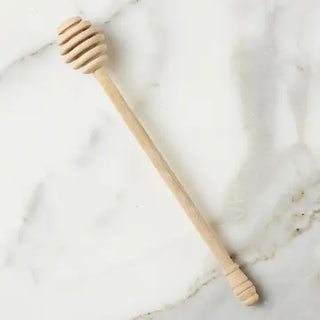 A light natural wood honey dipper with a ridged spoon head, placed diagonally on a white marble surface.
