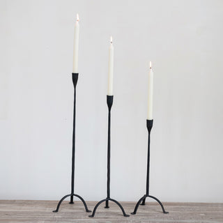Three black candle holders with white candles on a wooden floor against a gray wall.