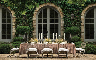 Outdoor dining setup with a table and chairs in front of a stone building with arched windows and greenery.