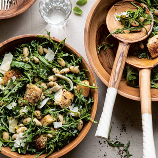 Wooden bowl filled with a green salad with croutons and beans, accompanied by a wooden spoon on a light background.