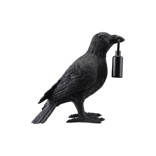 A black raven figurine holding a small black cylindrical bottle in its beak, shown against a plain white background.